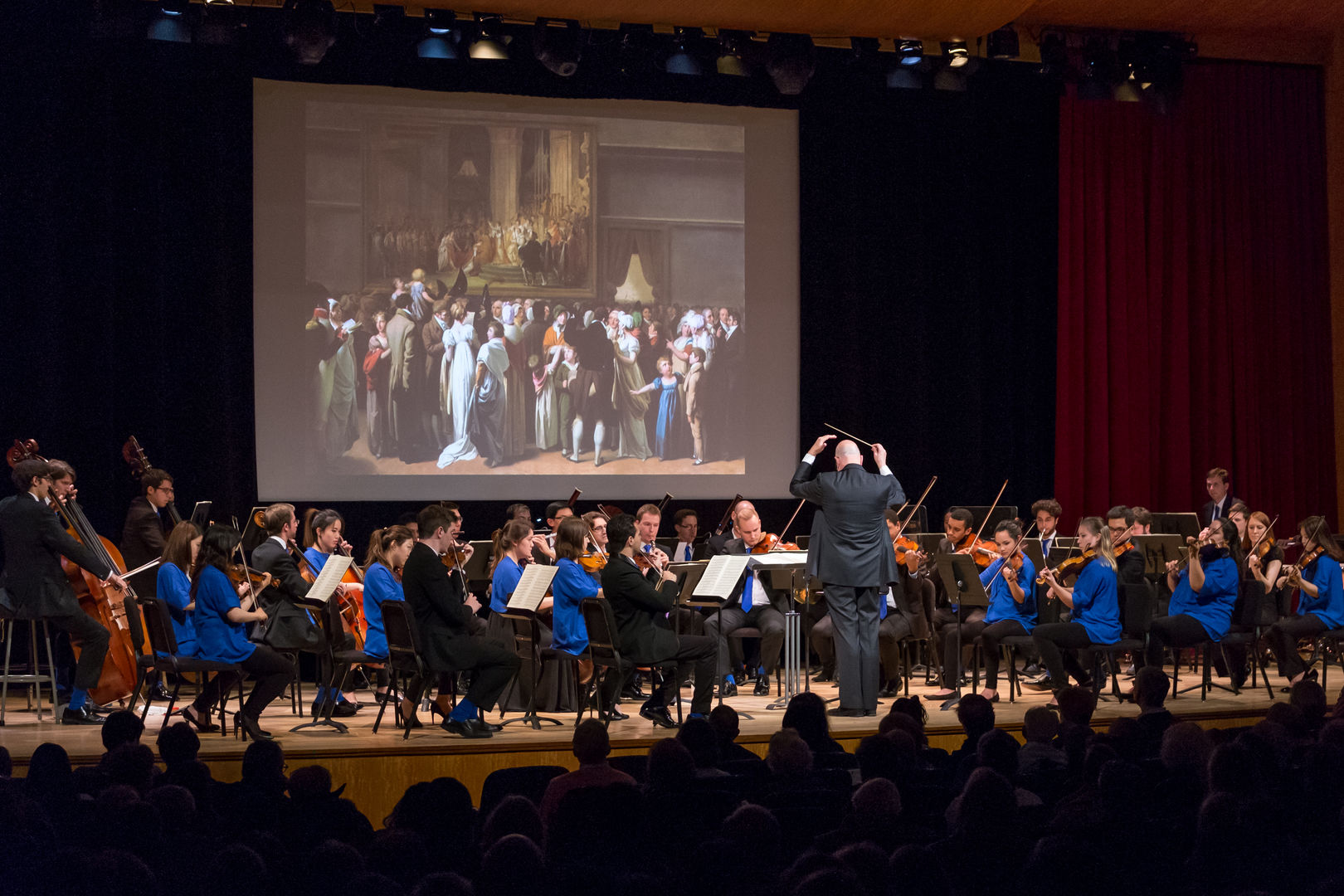 An orchestra clad in black and blue plays as a projection shows on a screen above.